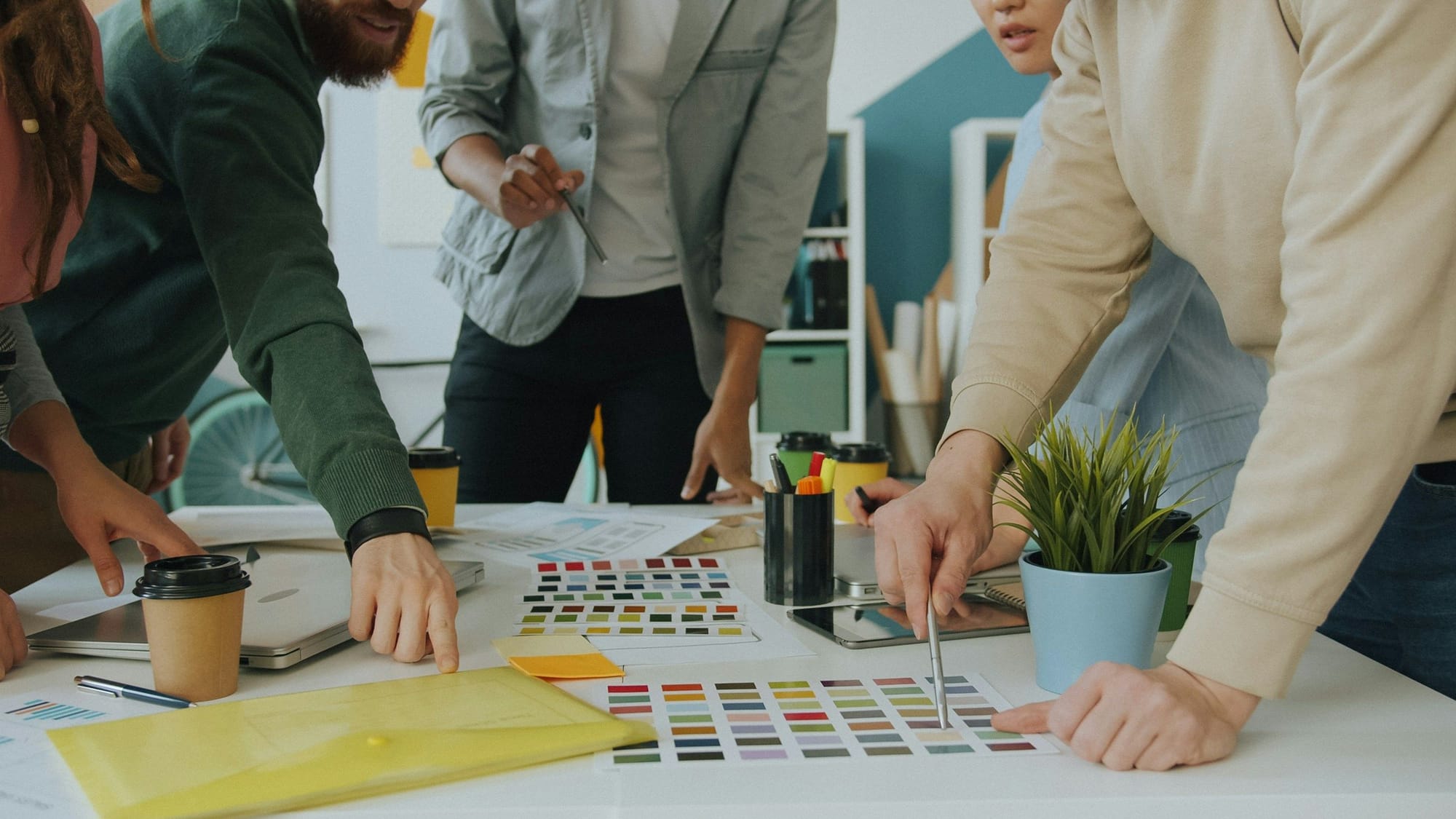 Team beugt sich über einen hellen Arbeitstisch mit Farbmustern, Skizzen und Kaffeebechern – Abstimmung im Designstudio
